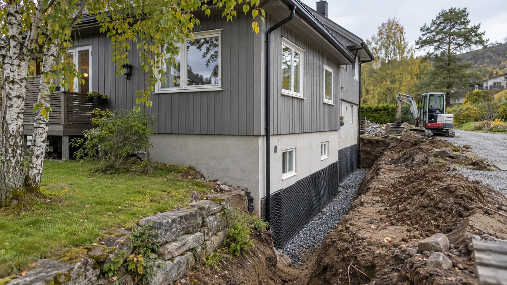 Dreneringsarbeid ved et norsk trehus, med åpen grøft og liten gravemaskin.